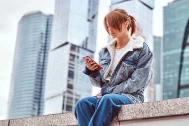 Yüzünde bulutlu sabah Moskova şehir içinde gökdelenler önünde oturan bir smartphone kullanarak denim ceket giyen dövmeli şık Kızıl saçlı hippi kız.