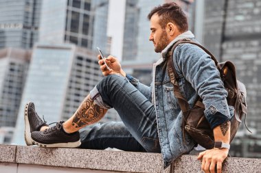 Dövmeli hipster adam bulutlu sabah Moskova şehir içinde gökdelenler önünde oturan smartphone kullanarak bir sırt çantası ile şık giyinmiş.