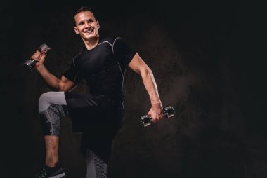 Koyu arka plan üzerinde dumbbells ile egzersiz yaparak spor giyim giyen genç bir vücut geliştirmeci.