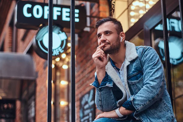 Een peinzende stijlvolle man draagt een denim jasje met draadloze hoofdtelefoons houden afhaalmaaltijden koffie buiten het café. — Stockfoto