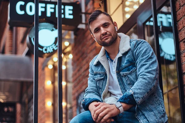 Een peinzende stijlvolle man draagt een denim jasje met draadloze hoofdtelefoons houden afhaalmaaltijden koffie buiten het café. — Stockfoto