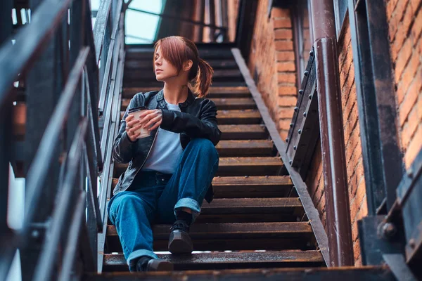 Uma menina bonita vestindo roupas da moda sentado nas escadas fora do café e segurando o café takeaway . — Fotografia de Stock