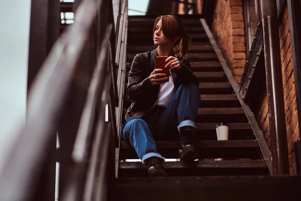 Красивая рыжая девушка в модной одежде сидит на лестнице возле кафе и держит смартфон . — стоковое фото