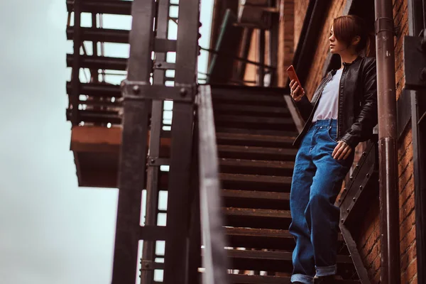 Una hermosa pelirroja con ropa de moda de pie en las escaleras fuera de la cafetería y sosteniendo un teléfono inteligente . — Foto de Stock