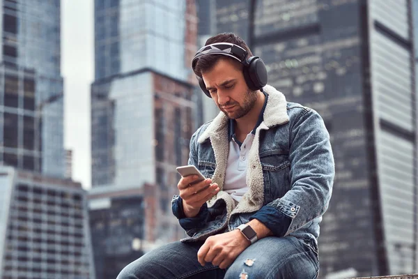 Elegante hombre con chaqueta de mezclilla escuchando música y usando un teléfono inteligente sentado frente a rascacielos en la ciudad de Moskow en la mañana nublada . — Foto de Stock