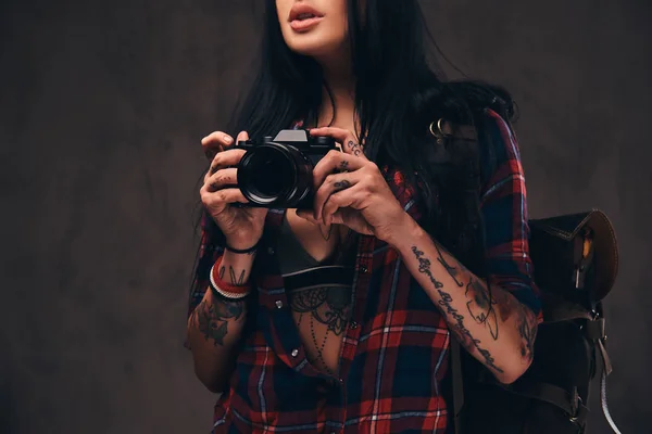 Tatuado hipster menina vestindo um vermelho desabotoado verificado camisa segurando uma câmera em um fundo escuro . — Fotografia de Stock