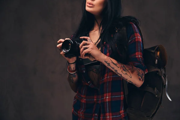 Tattooed hipster girl wearing a red unbuttoned checked shirt holding a camera on a dark background.