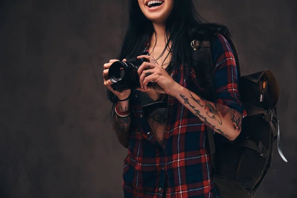 Tatuado hipster menina vestindo um vermelho desabotoado verificado camisa segurando uma câmera em um fundo escuro . — Fotografia de Stock