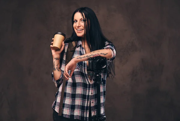 Getatoeëerd meisje houden een afhaalmaaltijden koffie terwijl leunend op een camera in een studio. — Stockfoto