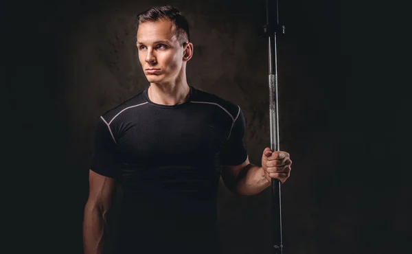Portret van een knappe jonge bodybuilder houden een barbell en weg op donkere achtergrond. — Stockfoto