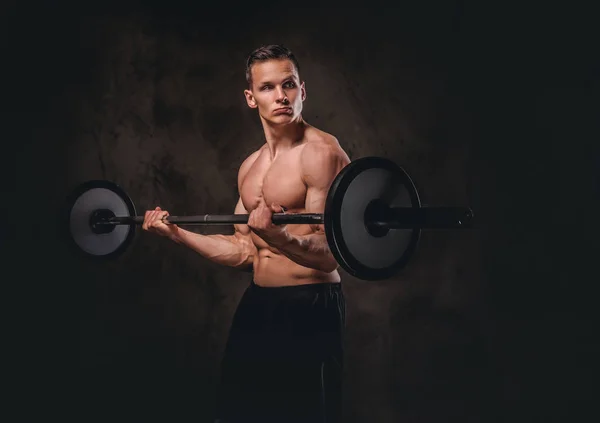 Jonge shirtless bodybuilder holding een barbell en doen oefening op de biceps. Geïsoleerd op donkere achtergrond. — Stockfoto