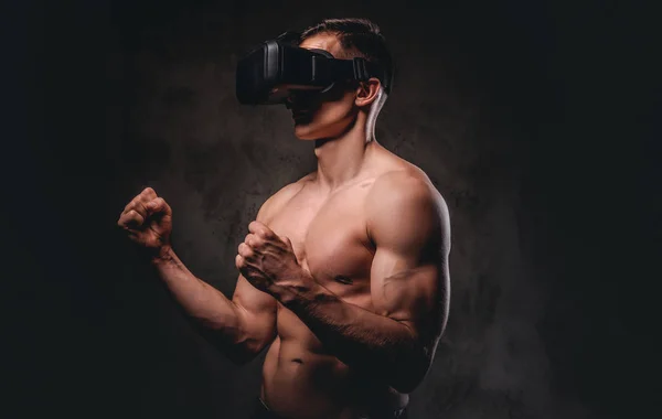 Young sportsman with muscular body wearing VR headset training punches in virtual reality fight on dark background.