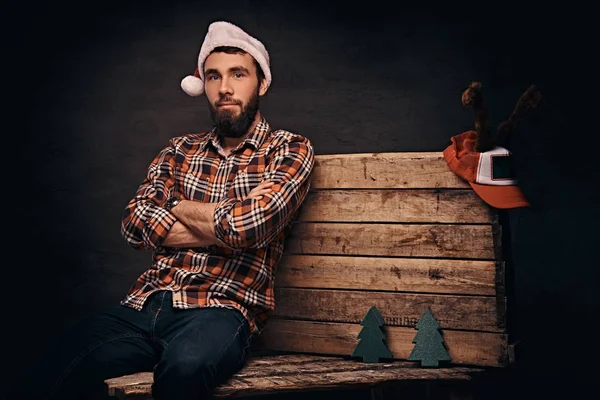 Ein bärtiger Mann mit Weihnachtsmütze sitzt mit verschränkten Armen auf einer Holzpalette. Weihnachtskonzept. — Stockfoto