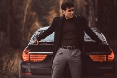 Acımasız bir genç adam bir lüks araba sonbahar ormandaki yaslanmış bir siyah palto giyiyor