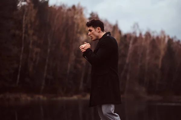 A handsome young man warms hands near a lake in the autumn forest. — Stock Photo, Image