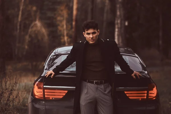 Un joven elegante con un abrigo negro apoyado en un coche de lujo en el bosque de otoño — Foto de Stock