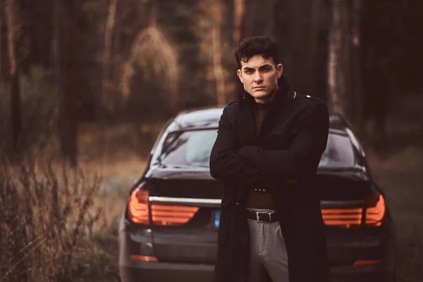 在秋天的森林里, 一个穿着黑色外套、胳膊交叉的时尚年轻人站在一辆豪华车旁边 — 图库照片