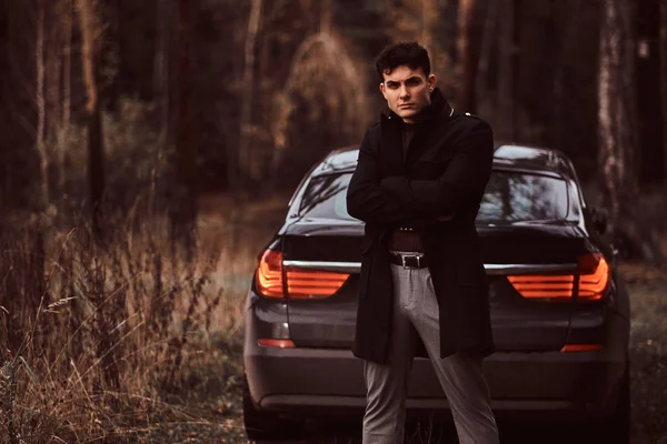 Un joven elegante con un abrigo negro con los brazos cruzados de pie junto a un coche de lujo en el bosque de otoño — Foto de Stock