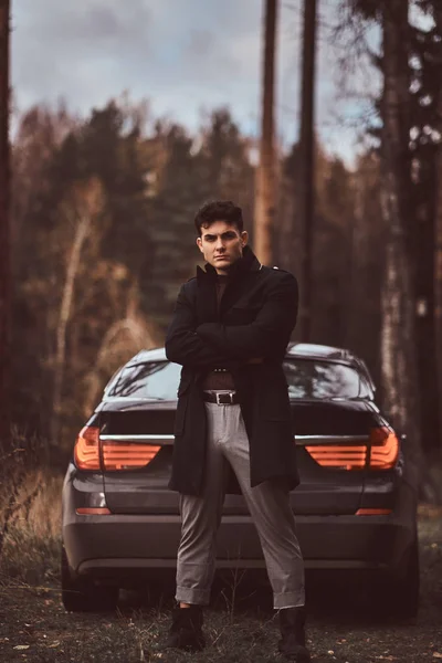 Un joven elegante con un abrigo negro con los brazos cruzados de pie junto a un coche de lujo en el bosque de otoño — Foto de Stock