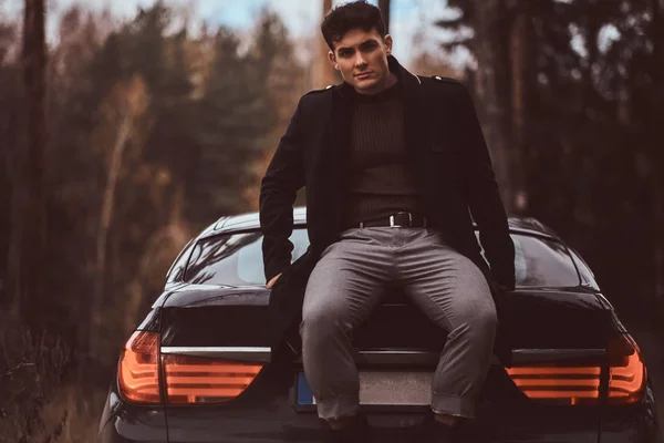 Un joven elegante con un abrigo negro sentado en el maletero de un coche en el bosque de otoño — Foto de Stock