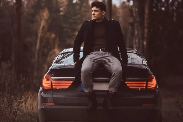 A stylish young man wearing a black coat sitting on the trunk of a car in the autumn forest — Stock Photo, Image