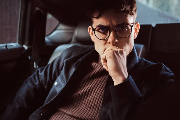 Un joven pensativo con gafas sentado en el asiento trasero del coche . — Foto de Stock