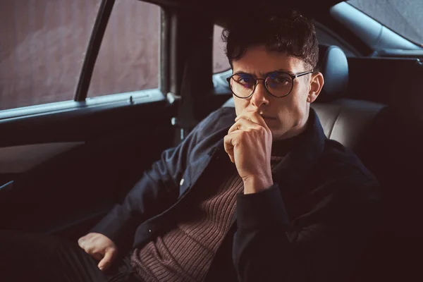 Un joven pensativo con gafas sentado en el asiento trasero del coche . — Foto de Stock
