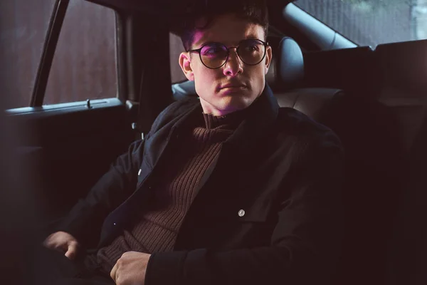 Un joven guapo con gafas sentado en el asiento trasero del coche . — Foto de Stock