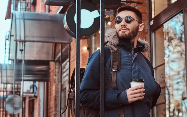 Un joven guapo con una mochila sosteniendo una taza de café mientras estaba parado cerca de un café afuera . —  Fotos de Stock