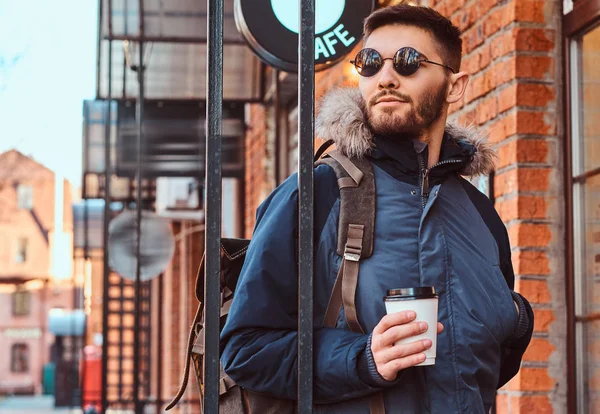 Un joven guapo con una mochila sosteniendo una taza de café mientras estaba parado cerca de un café afuera . —  Fotos de Stock