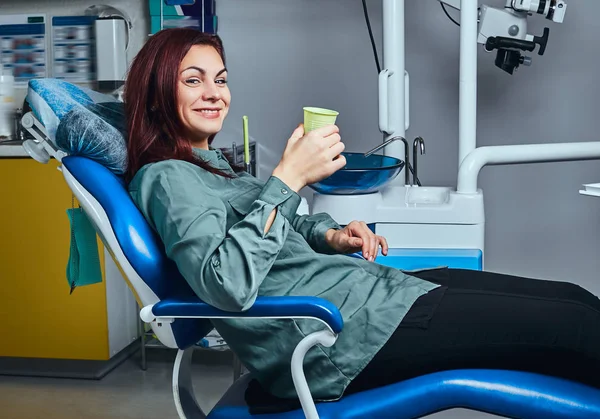 クリニックでマウスウォッシュでカップを保持している歯科医の椅子に座っている幸せな女. — ストック写真