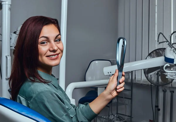 Schönes Mädchen sitzt im Zahnarztstuhl mit Spiegel in den Händen und blickt in die Kamera. — Stockfoto