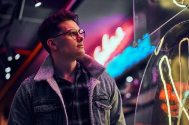 Genç bir adam sokakta geceleri ayakta modaya uygun giyinmiş. Işıklı tabela, neon, ışıklar.