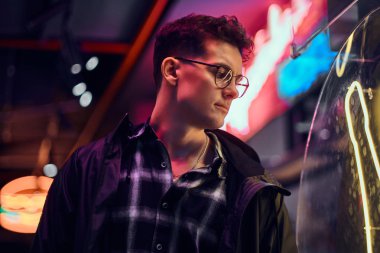 Geceleri sokağa ışıklı tabela bakarak spor kapüşonlu genç dalgın bir adam portresi.