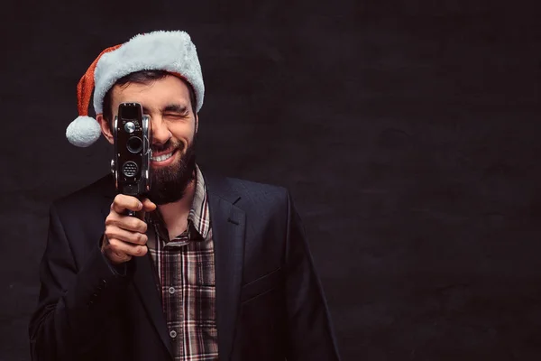 Estúdio retrato de homem barbudo vestindo um terno clássico e chapéu de Papai Noel segurando uma câmera retro, fazendo uma foto . — Fotografia de Stock