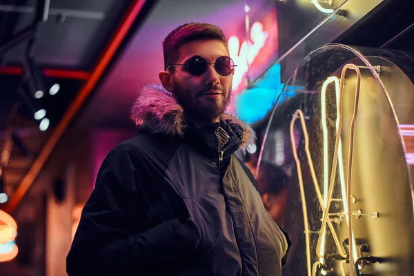 Un hombre guapo con un abrigo y gafas de sol con las manos en los bolsillos, de pie en la noche en la calle . — Foto de Stock