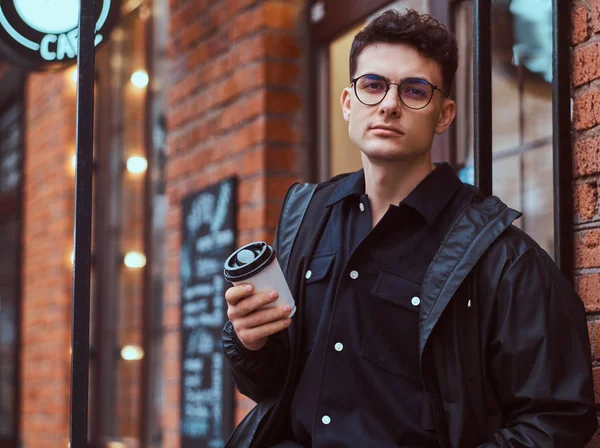 Un joven guapo sosteniendo un café para llevar mientras está parado cerca de un café afuera . — Foto de Stock