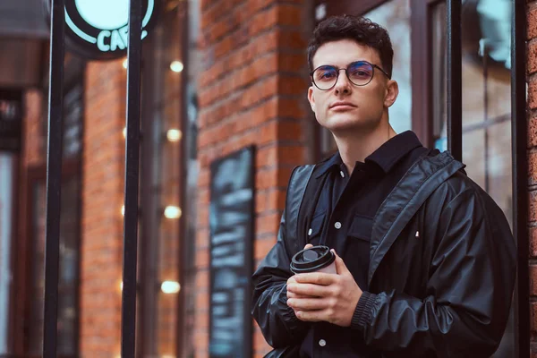 Un joven guapo sosteniendo un café para llevar mientras está parado cerca de un café afuera . — Foto de Stock