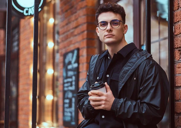 Un joven guapo sosteniendo un café para llevar mientras está parado cerca de un café afuera . — Foto de Stock