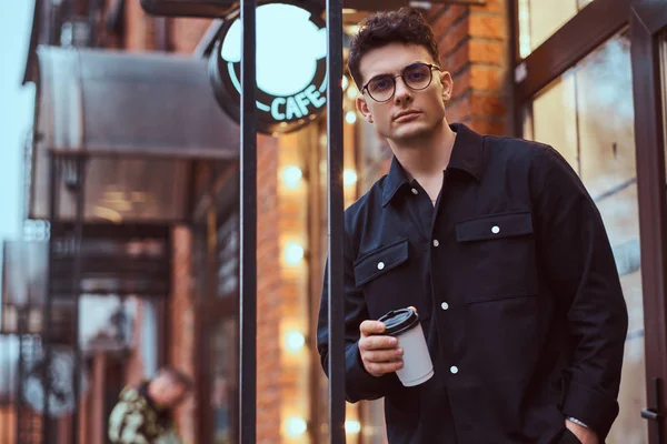 Un joven guapo sosteniendo un café para llevar mientras está parado cerca de un café afuera . — Foto de Stock