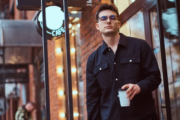 一个年轻的帅哥站在外面的咖啡馆附近拿着外卖咖啡. — 图库照片