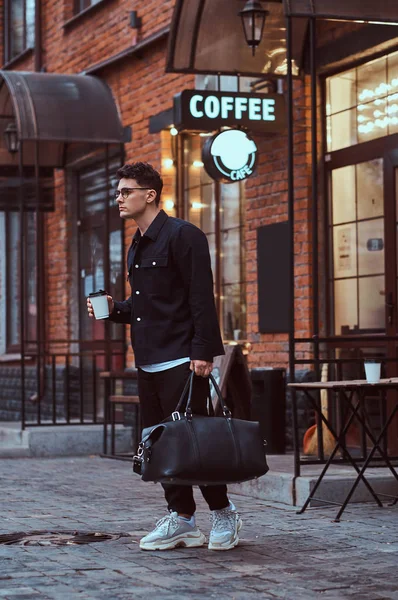 Een knappe man in een shirt holding een afhaalmaaltijden koffie en tas tijdens het wandelen in de straat voor brildragende. — Stockfoto