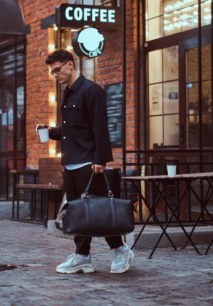 Egy jóképű férfi, szemüveget viselt ing gazdaság elvihető kávé és táska rövid idő gyaloglás legyőz a utca. — Stock Fotó