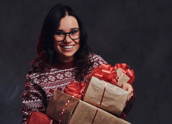 Portret van een gelukkig brunette meisje dragen van bril en warme trui houden een geschenken-dozen, geïsoleerd op een donkere gestructureerde achtergrond. — Stockfoto