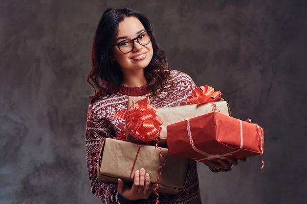 Retrato de uma menina morena feliz vestindo óculos e suéter quente segurando uma caixas de presentes, isolado em um fundo escuro texturizado . — Fotografia de Stock