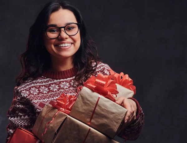 Retrato de uma menina morena feliz vestindo óculos e suéter quente segurando uma caixas de presentes, isolado em um fundo escuro texturizado . — Fotografia de Stock