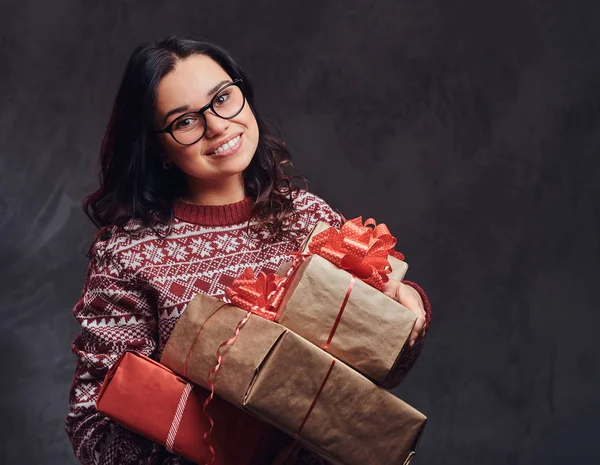 Portrait of a happy brunette girl wearing eyeglasses and warm sweater holding a gifts boxes, isolated on a dark textured background. — Stock Photo, Image