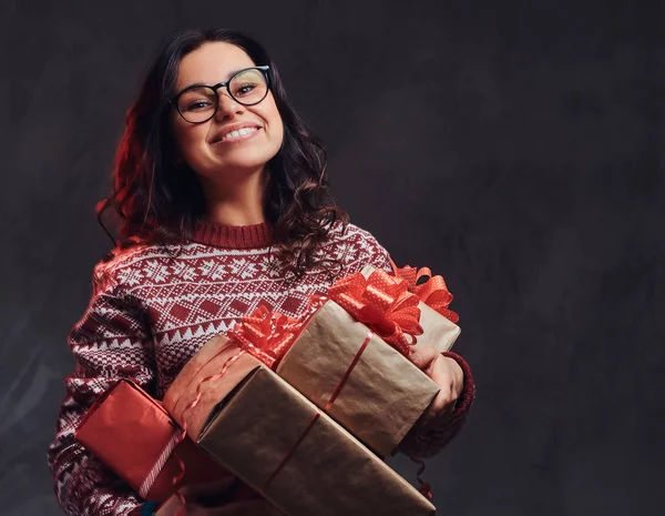 Retrato de uma menina morena feliz vestindo óculos e suéter quente segurando uma caixas de presentes, isolado em um fundo escuro texturizado . — Fotografia de Stock