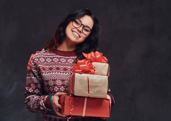 Πορτρέτο του ένα ευτυχισμένο κορίτσι μελαχρινή φορώντας γυαλιά και ζεστό πουλόβερ κρατώντας ένα κουτιά δώρων, απομονώνονται σε ένα σκοτεινό φόντο με υφή. — Φωτογραφία Αρχείου