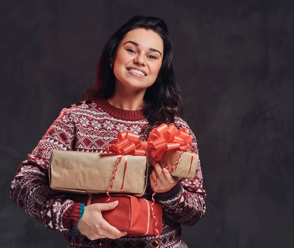 Retrato de uma menina morena feliz vestindo uma camisola quente segurando uma caixas de presentes, isolado em um fundo escuro texturizado . — Fotografia de Stock